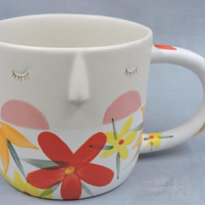 Puede incluir: Una taza de cer&aacute;mica con dise&ntilde;o de rostro, con ojos cerrados, nariz y patrones florales. Las flores son en tonos rojos, amarillos y naranjas. El asa tiene un dise&ntilde;o curvo y est&aacute; decorada con patrones florales similares.