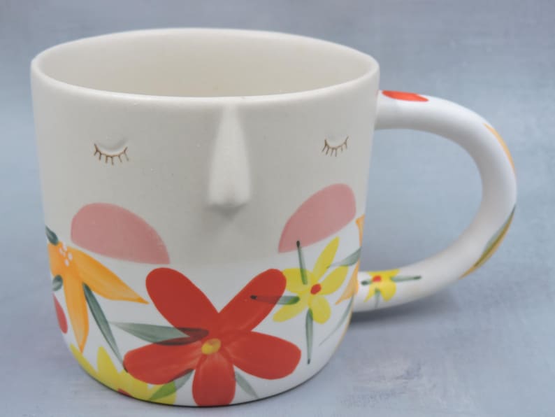 Puede incluir: Una taza de cer&aacute;mica con dise&ntilde;o de rostro, con ojos cerrados, nariz y patrones florales. Las flores son en tonos rojos, amarillos y naranjas. El asa tiene un dise&ntilde;o curvo y est&aacute; decorada con patrones florales similares.