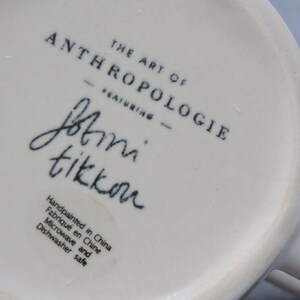 Puede incluir: Primer plano de la base de una taza de cer&aacute;mica blanca. El texto "THE ART OF ANTHROPOLOGIE" est&aacute; impreso en negro, junto con la firma "Jbtini tikkou". La taza est&aacute; pintada a mano en China y es apta para microondas y lavavajillas.