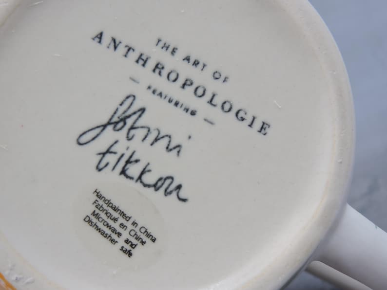 Puede incluir: Primer plano de la base de una taza de cer&aacute;mica blanca. El texto "THE ART OF ANTHROPOLOGIE" est&aacute; impreso en negro, junto con la firma "Jbtini tikkou". La taza est&aacute; pintada a mano en China y es apta para microondas y lavavajillas.