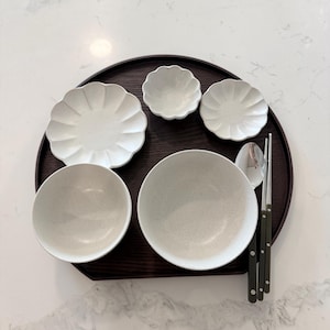 May include: A dark brown circular wooden tray holding two speckled off-white bowls, two scalloped-edge plates, one small scalloped bowl, a silver spoon, and a pair of dark brown chopsticks with silver tips arranged in a neat dining set.