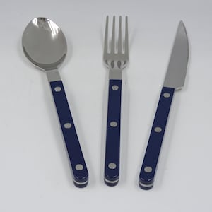 May include: A stainless steel spoon, fork, and knife set. The utensils have navy blue handles with three silver rivets. The set is designed for dining and is placed on a white surface.