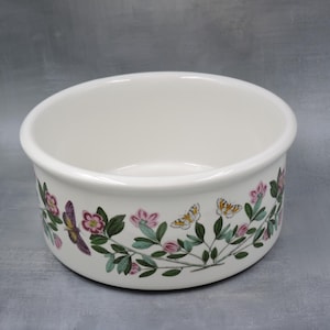 May include: A white ceramic bowl with a floral and butterfly design. The bowl features a wide rim and a decorative band with pink flowers, green leaves, and butterflies in shades of purple and yellow.