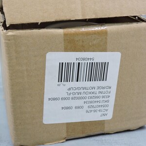 Puede incluir: Una caja de cart&oacute;n marr&oacute;n con una etiqueta blanca y un c&oacute;digo de barras. La etiqueta incluye el texto "FOTINI TIKKOU MUG-FL" y "RD/RGE MOTMUG/CUP". La caja est&aacute; parcialmente abierta, revelando el contenido. La caja est&aacute; sellada con cinta adhesiva marr&oacute;n.