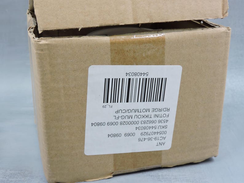 Puede incluir: Una caja de cart&oacute;n marr&oacute;n con una etiqueta blanca y un c&oacute;digo de barras. La etiqueta incluye el texto "FOTINI TIKKOU MUG-FL" y "RD/RGE MOTMUG/CUP". La caja est&aacute; parcialmente abierta, revelando el contenido. La caja est&aacute; sellada con cinta adhesiva marr&oacute;n.