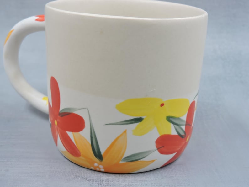 Puede incluir: Taza de cer&aacute;mica color crema con un dise&ntilde;o floral pintado a mano en rojo, amarillo y naranja, con hojas verdes. El asa tambi&eacute;n est&aacute; decorada con flores. Una taza decorativa para bebidas calientes.