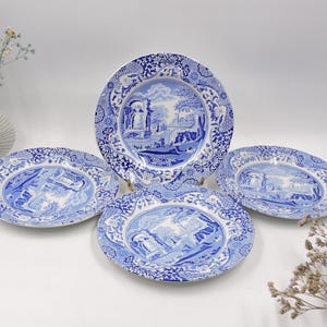 May include: A set of four blue and white ceramic plates. The plates feature a detailed landscape design with architectural elements. The plates are arranged on a white surface, with a vase of flowers in the background.
