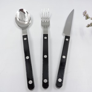 May include: A stainless steel spoon, fork, and knife set with black handles. The utensils have a classic design, with a polished finish on the spoon and knife blade. The black handles are accented with three silver rivets each. The set is ideal for everyday dining.