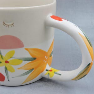 Puede incluir: Taza de cer&aacute;mica blanca con dise&ntilde;o floral. La taza presenta una forma de sol rosa, flores amarillas y naranjas y un asa pintada. El asa est&aacute; decorada con detalles rojos, verdes y amarillos. Un peque&ntilde;o dise&ntilde;o de pesta&ntilde;as est&aacute; pintado en el lateral.