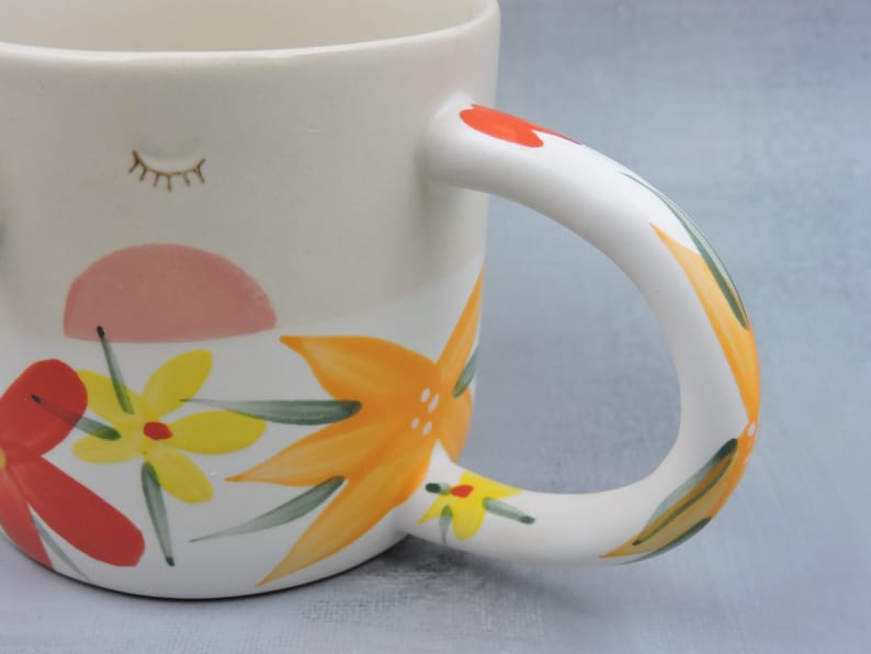 Puede incluir: Taza de cer&aacute;mica blanca con dise&ntilde;o floral. La taza presenta una forma de sol rosa, flores amarillas y naranjas y un asa pintada. El asa est&aacute; decorada con detalles rojos, verdes y amarillos. Un peque&ntilde;o dise&ntilde;o de pesta&ntilde;as est&aacute; pintado en el lateral.
