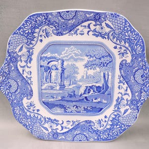 May include: Square blue and white decorative plate with a landscape scene in the center. The plate has a scalloped edge and floral patterns. The central image depicts a classical landscape with buildings, trees, and figures.