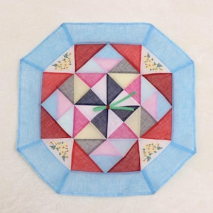 May include: An octagonal wall clock with a patchwork design. The clock face features a geometric pattern of red, pink, blue, gray, and white triangles. The clock hands are green. The clock is surrounded by a light blue border with floral accents.