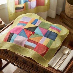May include: A square, multi-colored patchwork fabric cover featuring a geometric pattern of triangles and rectangles in shades of red, blue, yellow, green, and purple, with a light green border and a small red decorative knot in the center.