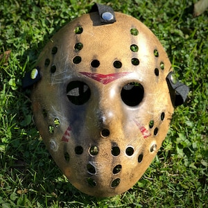 Peut inclure: Un masque de hockey beige vieilli avec une finition usée et des ombres brun foncé, de nombreux trous de ventilation circulaires et trois marques triangulaires rouges distinctes au-dessus des yeux et sur les joues, fixé par des sangles élastiques noires.