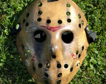 May include: A weathered tan hockey mask featuring a distressed finish with dark brown shading, numerous circular ventilation holes, and three distinct red triangular markings above the eyes and on the cheeks, secured by black elastic straps.
