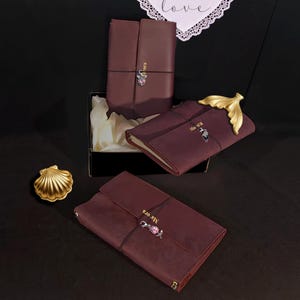 Può includere: Diari in pelle bordeaux con chiusure decorative e la parola "Ma ora" stampata sul davanti. Un diario ha una chiusura a coda di pesce dorata. Una conchiglia dorata è a sinistra. I diari sono in una scatola nera con carta velina bianca.