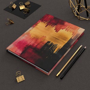 Könnte beinhalten: Ein Hardcover-Journal mit einem abstrakten Design in Rot-, Orange- und Schwarztönen. Gold- und schwarze Bleistifte und goldene Büroklammern sind um das Journal herum auf einer dunkelgrauen Oberfläche angeordnet.