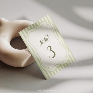 May include: A light green and white striped table number card with the number 3. The card has a decorative border and the word "table" in script. A beige, abstract sculpture is in the background.