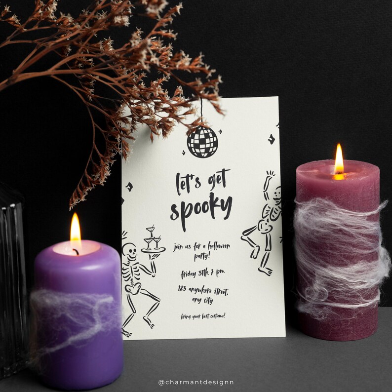 May include: A Halloween party invitation with the text "Let's get spooky" and skeleton illustrations. Two lit pillar candles, one purple and one burgundy, are decorated with faux spiderwebs. Dried floral accents complete the festive scene.