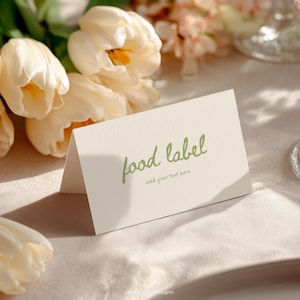 Può includere: Un biglietto piegato, bianco sporco, con le parole "food label" in verde. Il biglietto è su una tovaglia beige, con tulipani bianchi e altri fiori sullo sfondo. Il testo "add your text here" è sotto.