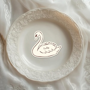 May include: A sticker featuring a swan design with the name "Hailey" in cursive script. The sticker is cream-colored with a dark brown outline and details. It is placed on a white plate with a floral embossed border, set against a white fabric background.