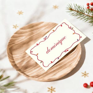 Puede incluir: Una tarjeta de sitio crema con el nombre "Dominique" en escritura roja, enmarcada por una guirnalda de luces roja. La tarjeta descansa sobre un plato de madera, con decoraciones festivas en el fondo.