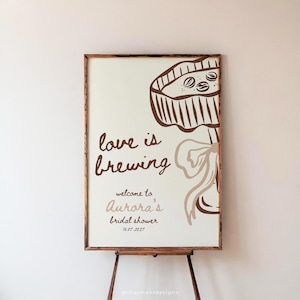 以下が含まれることがあります： 「love is brewing」という茶色の筆記体で書かれた額入りの看板。11.07.2027に「Aurora's bridal shower」へようこそ。コーヒーポットとリボンの茶色のイラストが右上を飾っています。看板は木製のイーゼルに表示されています。