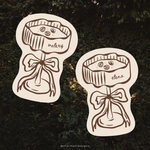 Könnte beinhalten: Zwei braune Illustrationen eines Kuchens auf einem Ständer mit einer Schleife. Ein Kuchen trägt den Namen "valery" und der andere "elena". Die Kuchen haben drei Kaffeebohnen oben drauf. Der Hintergrund ist dunkelgrün.