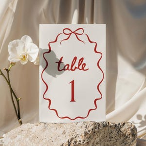 Op de afbeelding: Een witte tafelnummerkaart met rode tekst "table 1". De kaart is omlijst door een rode golvende rand met een strik aan de bovenkant. Een witte orchidee en een steen zijn op de achtergrond.