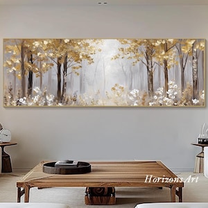 Puede incluir: Una pintura larga y horizontal que representa una escena de bosque con hojas doradas y flores blancas. La pintura está enmarcada en un marco fino de oro y cuelga de una pared blanca sobre una mesa de café de madera.