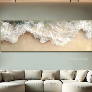May include: A horizontal abstract painting of ocean waves crashing on a sandy beach. The painting is in shades of white, beige, and blue, with a gold frame.