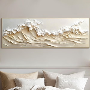 May include: A white and beige textured canvas wall art depicting a wave pattern. The art is framed in a thin gold frame and is hanging on a white wall above a bed with white bedding and a tan throw blanket.