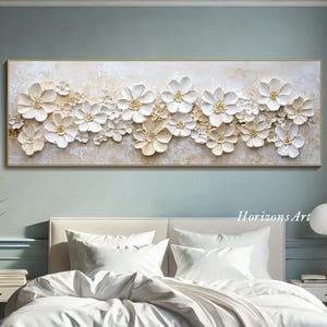 K&ouml;nnte beinhalten: Ein gro&szlig;er wei&szlig;er und goldener floralem Leinwand-Wandkunstdruck mit 3D-Effekt. Der Druck zeigt eine Vielzahl von wei&szlig;en Blumen mit goldenen Akzenten auf wei&szlig;em Hintergrund. Der Druck h&auml;ngt an einer Wand &uuml;ber einem wei&szlig;en Bett mit wei&szlig;en Bettw&auml;sche.