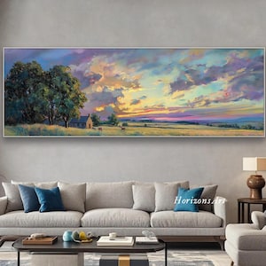 May include: A landscape painting depicts a vibrant sunset over a field with a small house and trees. The sky is filled with colorful clouds, and the artwork is framed in a silver border. The painting is displayed above a gray sofa.