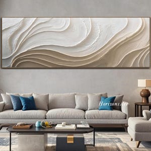 Könnte beinhalten: Ein großes, rechteckiges abstraktes Wandkunstwerk mit einem strukturierten, geschichteten Design in Weiß- und Beigetönen. Das Kunstwerk ist in einem dünnen, goldfarbenen Rahmen eingefasst. Das Stück wird über einem grauen Sofa in einem Wohnzimmer präsentiert.