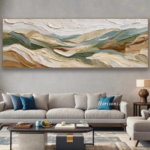 Puede incluir: Pintura de paisaje texturizada en un marco dorado. La obra de arte presenta una representación en capas de colinas onduladas en tonos de blanco, verde y marrón. La pintura se exhibe sobre un sofá gris en un salón.
