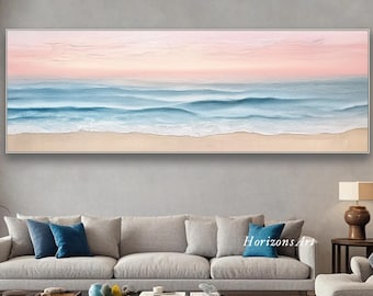 Puede incluir: Una pintura rectangular enmarcada representa una serena escena de playa. La obra presenta un cielo rosa y azul pastel, con olas de océano azul claro que rompen en una playa arenosa. La pintura se exhibe sobre un sofá gris.