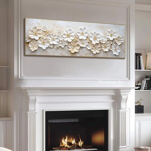K&ouml;nnte beinhalten: Ein gro&szlig;es, rechteckiges Leinwandbild mit einem wei&szlig;en und goldenen Blumendesign. Die Blumen sind erhaben und strukturiert, was dem Bild einen 3D-Effekt verleiht. Das Bild h&auml;ngt &uuml;ber einem wei&szlig;en Kamin.