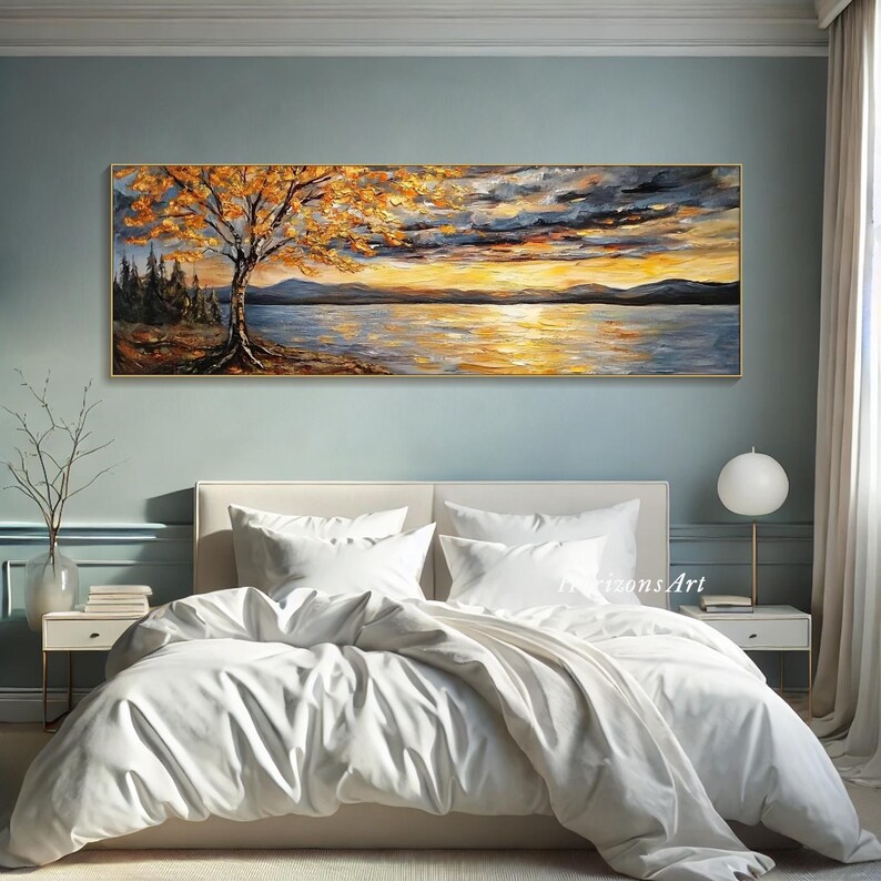 May include: A painting of a lake scene with a tree in the foreground and a sunset in the background. The painting is done in a palette of warm colors, including yellow, orange, and red. The painting is displayed on a wall above a white bed with white bedding.