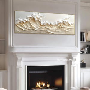 May include: A white and gold textured wall art piece depicting a wave crashing. The art is framed in a thin gold frame and is hanging above a white fireplace.