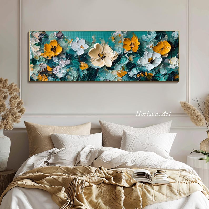 May include: Abstract painting featuring a floral design with white and yellow flowers on a teal background. The painting is framed in a gold frame and is hanging on a white wall above a bed with white bedding and a brown throw blanket.