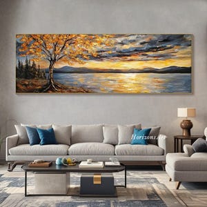 May include: A long, horizontal painting depicting a lake scene with a tree in the foreground. The tree has yellow and orange leaves. The lake is a deep blue with a golden sunset reflecting on the water. The sky is a mix of gray and blue with clouds. The painting is framed in a gold frame.