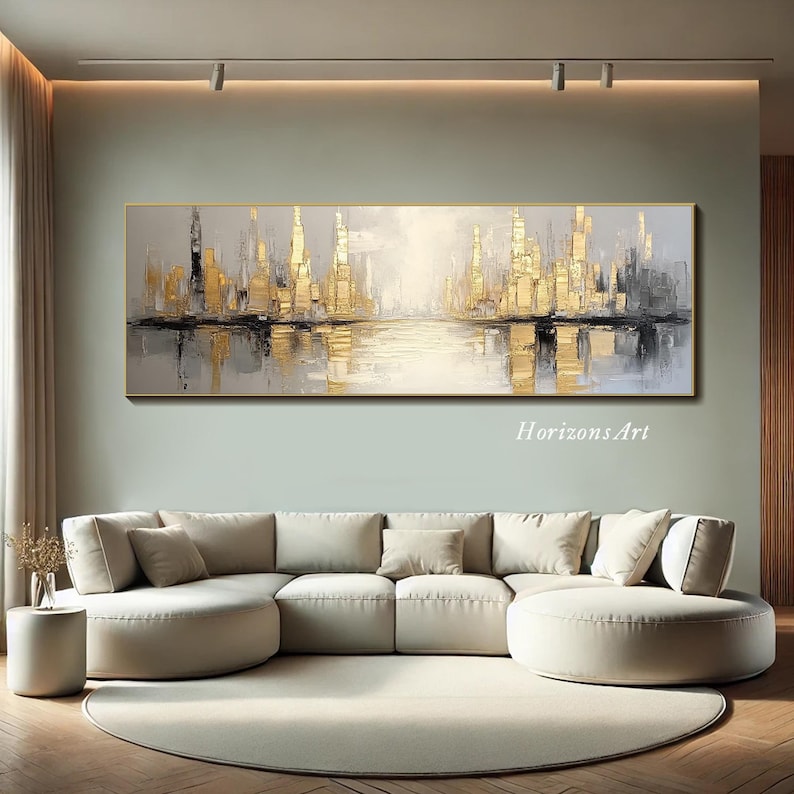 Puede incluir: Una gran pintura abstracta de una ciudad con tonos dorados y grises. La pintura representa un horizonte urbano con edificios reflejados en el agua. La pintura est&aacute; colgada en una pared en una sala de estar con un sof&aacute; blanco.