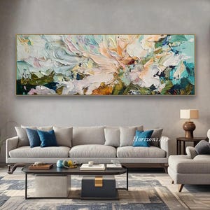 May include: Abstract painting with a gold frame, featuring a colorful, textured design with shades of blue, green, pink, and white. The painting is displayed on a wall above a gray sofa and a coffee table.