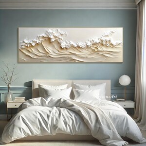 May include: A white and beige textured wall art piece depicting a wave pattern. The art is hanging above a white bed with white bedding.