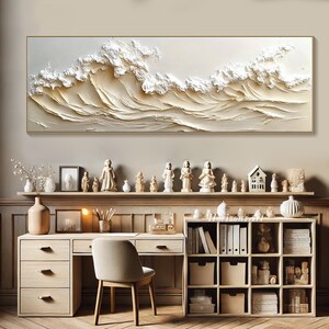 May include: A white and gold abstract painting of waves, framed in a thin gold frame, hangs above a wooden desk with a white chair. The desk has a shelf with books and a drawer. The wall is a light beige color.