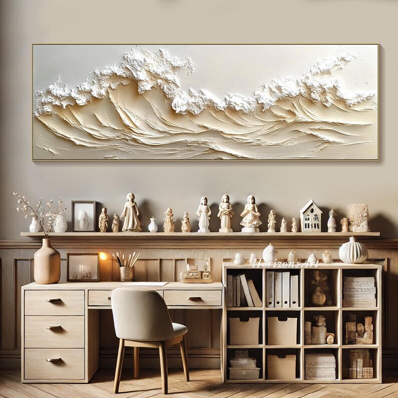 May include: A white and gold abstract painting of waves, framed in a thin gold frame, hangs above a wooden desk with a white chair. The desk has a shelf with books and a drawer. The wall is a light beige color.