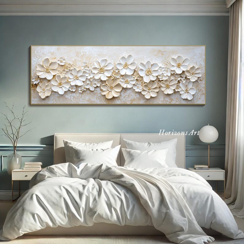 K&ouml;nnte beinhalten: Ein gro&szlig;er wei&szlig;er und goldener floralem Leinwand-Wandkunstdruck mit 3D-Effekt. Der Druck zeigt eine Vielzahl von wei&szlig;en Blumen mit goldenen Akzenten auf wei&szlig;em Hintergrund. Der Druck h&auml;ngt an einer Wand &uuml;ber einem wei&szlig;en Bett mit wei&szlig;en Bettw&auml;sche.