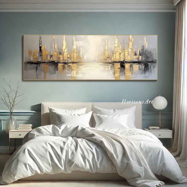 Puede incluir: Pintura abstracta de un paisaje urbano con tonos dorados y grises. La pintura representa un horizonte urbano con edificios reflejados en una masa de agua. La pintura est&aacute; enmarcada y colgada en una pared sobre una cama blanca con ropa de cama blanca.