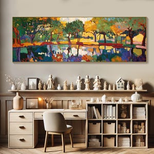 May include: A colorful abstract painting of a forest scene with trees reflected in a still body of water. The painting is framed in a gold frame and hangs on a wall above a desk and bookshelf.